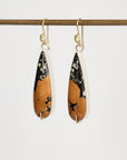 A pair of teardrop-shaped dangle earrings with a black and orange abstract pattern, suspended from gold fishhook wires and hanging from a gold rod against a plain light background.