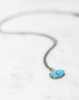 A delicate necklace with a small oval turquoise pendant in a gold-toned bezel setting, suspended from a thin dark chain on a light marble surface.