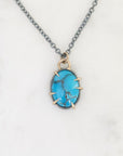 A turquoise stone pendant necklace with a silver-toned chain and gold-toned prong setting, displayed on a white marble surface.