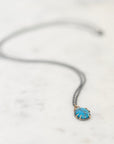 A delicate dark gray chain necklace with a small oval pendant featuring a bright turquoise blue center and gold-toned edges, resting on a light marble surface.