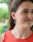 A close-up portrait of a woman wearing a dangling earring with a blue and white patterned stone or bead, photographed outdoors with a blurred green background.