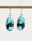 A pair of oval drop earrings with bright turquoise blue and black marbled stones, held in gold-toned prong settings with gold hooks, hanging from a gold rod against a light gray background.