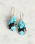 A pair of drop earrings with oval turquoise and black marbled stones in silver wire wrap, hanging from gold-tone fishhook wires on a light marble surface.