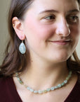 A woman wearing a light blue-green beaded necklace and matching teardrop earrings with gold-toned settings, shown in a close-up portrait.