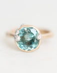 A round bezel-set ring featuring a large light blue gemstone in a gold-toned setting, accented with small clear stones on the shoulders, shown on a plain light background.