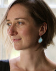 A woman with short light brown hair is shown in profile wearing a dangling earring with a light blue oval stone in a silver-toned setting.