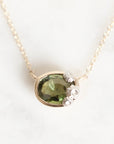 A close-up of a gold necklace with a pendant featuring a faceted olive green gemstone and small clear stones, shown on a light marble-like background.