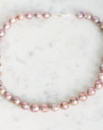 A strand of irregular baroque pearls in a dusty pink color, arranged in a circle on a white marble surface.