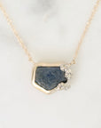 A gold necklace with a raw, irregular dark blue gemstone pendant and small clear crystals on one corner, displayed on a light marble surface.