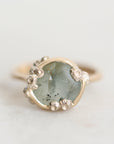 A close-up shot of a ring featuring a round, pale green stone with dark inclusions, surrounded by a gold-toned bezel and small silver-toned accents, set against a light off-white background.