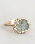 A close-up shot of a gold ring featuring a large, irregular pale green stone with tiny clear accent stones around its edge, set against a soft, light gray background.