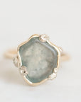 A close-up shot of a raw, shield-shaped light blue-green gemstone in a gold and silver floral bezel setting, on a plain white background.