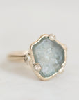 A gold ring featuring a large, irregular-shaped light blue-gray druzy stone with tiny inclusions, accented by three small clear crystals along the band, set against a clean white background.