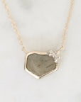 A delicate gold chain necklace featuring an irregular, raw stone pendant with a small cluster of clear crystals at the top right, displayed on a light marble surface.