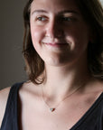 A close-up portrait of a woman wearing a delicate gold chain necklace with a small, round, light-colored stone pendant, set against a soft-focus indoor background.
