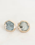 A pair of gold stud earrings featuring irregular, polished light blue stone slices with tiny clear accent stones along the edges, set against a plain white background.