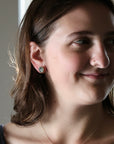 A close-up portrait of a woman with brown hair wearing a small round stud earring with a greenish-gray stone, shown in profile against a softly lit indoor background.