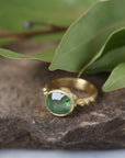 Gold ring with a green gemstone on a stone surface with leaves in the background