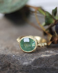 Gold ring with a green gemstone on a stone surface with leaves in the background