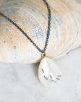 A close-up of a necklace with a dark chain and a teardrop-shaped white pendant featuring small fish designs, resting on a seashell over a light marble surface.