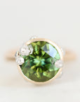 A round-cut green gemstone ring set in a gold-toned bezel with small clear accent stones, photographed on a clean light gray background.