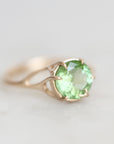 A gold ring featuring a round-cut light green gemstone in a prong setting, shown on a soft, light gray background.