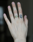 A close-up of a person's hand wearing a gold ring with a green oval gemstone, with a small black geometric tattoo on the side of the hand.
