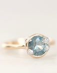 A gold ring featuring a round-cut light blue gemstone in a prong setting, accented with small clear stones on the shoulders, shown against a plain light background.