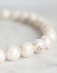 A close-up of a pearl beaded bracelet on a light marble surface, showing glossy white pearls and a small silver-toned spacer bead.