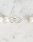 A close-up of a pearl necklace with a silver fishhook clasp on a light marble surface.