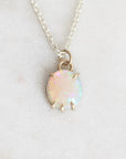 A delicate silver chain necklace with an oval opal pendant in a gold-toned prong setting, resting on a light gray textured surface.