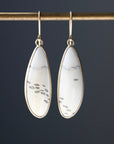 A pair of gold drop earrings with teardrop-shaped white stone inlays featuring small black fish motifs, hanging from a gold rod against a dark gray background.