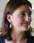 A woman with shoulder-length brown hair wearing a small white teardrop dangle earring, shown in a close-up portrait against a softly blurred indoor background.