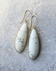 A pair of gold drop earrings with teardrop-shaped white stone inlays featuring black veining, displayed on a textured gray surface.