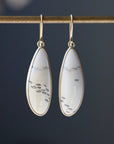 A pair of gold drop earrings with teardrop-shaped white stone pendants featuring black fish motifs, hanging from a gold rod against a dark gray background.
