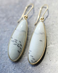 A pair of teardrop-shaped dangle earrings with white marbled stone inlays featuring small black fish motifs, set in gold-toned metal with matching gold fishhook ear wires, displayed on a textured gray surface.