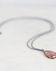 A delicate dark gray chain necklace with a small oval pendant featuring a pink stone in a gold-toned bezel setting, resting on a light off-white surface.