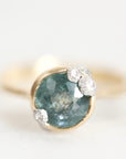 A gold ring featuring a large oval faceted blue-green gemstone with three small clear round-cut accent stones clustered at the top right, photographed on a clean white background.