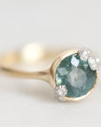 A gold ring featuring a round faceted teal-blue gemstone in a prong setting, accented with three small clear diamonds, photographed on a soft light gray background.