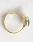 A gold ring with a small dark green gemstone and clear accent stones, photographed on a light marble surface.