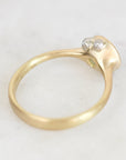 A close-up of a yellow gold ring with a textured, organic-shaped bezel setting holding two small clear gemstones, photographed on a light marble surface.