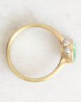 A gold ring with a bezel-set light green oval gemstone and two small clear stones, photographed on a light marble-like surface.
