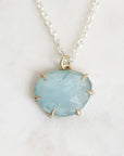 A light blue stone pendant necklace with a silver chain and a gold-toned prong setting, displayed on a light marble surface.