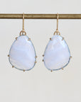 A pair of teardrop-shaped drop earrings with pale iridescent blue stones in gold prong settings and hooks, hanging from a gold rod against a light gray background.