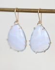 A pair of teardrop-shaped light blue gemstone drop earrings with gold hooks and bezel settings, hanging from a gold-colored horizontal bar against a light gray background.