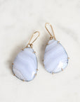 A pair of teardrop-shaped drop earrings featuring pale blue and white agate stone pendants with gold wire-wrapped edges and gold fishhook ear wires, displayed on a light marble surface.