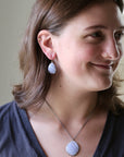 A woman wearing a matching light blue and white seashell pendant necklace and matching drop earrings, shown in a close-up portrait.