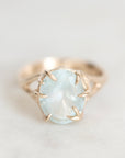 A gold ring featuring a large, oval-cut light blue gemstone in a prong setting, shown on a soft, light gray background.
