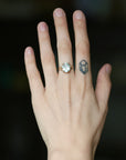 A close-up of a person's hand wearing a gold ring with a round white stone, with a small geometric tattoo on the middle finger, against a dark blurred background.