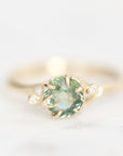 A yellow gold ring featuring a round green gemstone in a prong setting, accented by two small round clear stones on the shoulders, set against a soft white background.